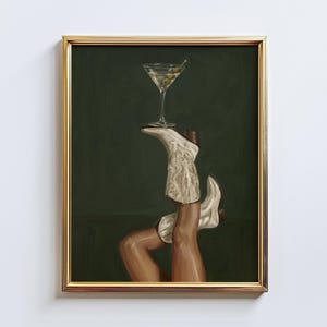 Op de afbeelding: Een ingelijste kunstdruk met een martini-glas in evenwicht op een paar witte cowboylaarzen. De laarzen zijn versierd met decoratieve stiksels. De achtergrond is donkergroen en de lijst is goudkleurig.
