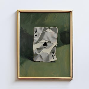 Op de afbeelding: Een ingelijst schilderij van een gekreukelde Aas van Schoppen speelkaart. De kaart is wit met zwarte schoppensymbolen en de letter "A" in elke hoek. De achtergrond is een groene, getextureerde ondergrond.
