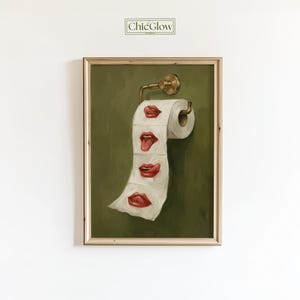 Op de afbeelding: Een ingelijst kunstwerk met een rol toiletpapier met geschilderde rode lippen in verschillende uitdrukkingen. Het kunstwerk is geplaatst tegen een groene achtergrond en heeft een goudkleurige toiletpapierhouder. Het kunstwerk is gelabeld "ChicGlow".