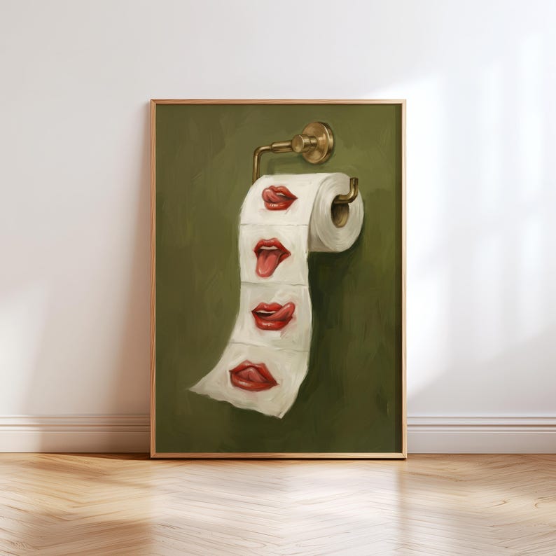 Op de afbeelding: Een ingelijste kunstdruk met een rol toiletpapier met geschilderde rode lippen op elk vel. Het kunstwerk is gemonteerd op een messing toiletpapierhouder tegen een groene achtergrond. De lijst is goudkleurig.