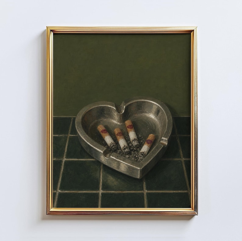 May include: A framed wall art print featuring a heart-shaped metallic ashtray resting on a dark green tiled surface. Inside the ashtray are four cigarette butts, each marked with a small red lipstick kiss print on the white filter section.