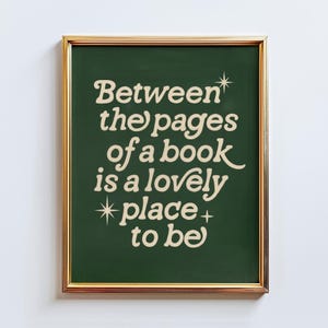 Può includere: Una stampa incorniciata con il testo "Between the pages of a book is a lovely place to be" in un carattere stravagante color crema. Il testo è su uno sfondo verde scuro, incorniciato da un bordo dorato.