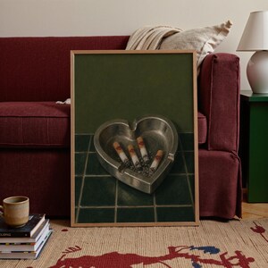 May include: A framed art print featuring a heart-shaped metallic ashtray resting on dark green square tiles. Inside the ashtray are four cigarette butts, each marked with a small red lipstick kiss print on the filter, set against a dark green wall.
