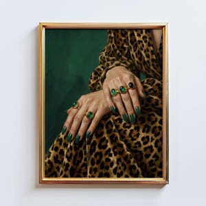 May include: A framed print featuring hands adorned with emerald green rings and matching nail polish. The person is wearing a leopard print garment. The background is a deep green color, and the frame is gold.