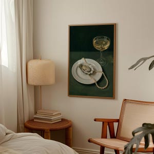 May include: A framed painting depicts a still life with a glass of wine, a white plate with an oyster, and a pearl necklace. The background is a dark green, and the painting is in a wooden frame. The style is reminiscent of classic still life paintings.