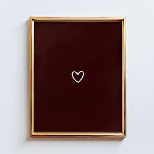 May include: A framed art print featuring a gold-colored frame and a dark burgundy background. A white outline of a heart is centered on the burgundy background. The artwork is displayed against a white wall.