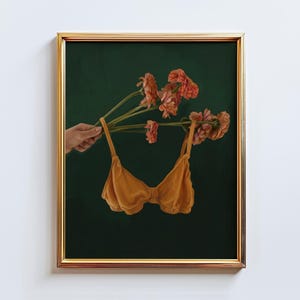 May include: A framed art print featuring a mustard-yellow bra suspended from a bouquet of peach-colored flowers against a dark green background. The artwork is in a gold frame, creating a striking contrast.
