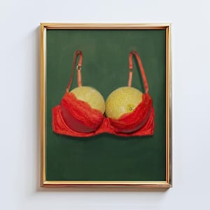 May include: A framed art print featuring a red bra with lace detailing, holding two cantaloupes. The artwork is set against a dark green background and framed in gold.