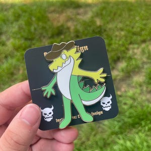 Puede incluir: Un pin de esmalte verde, amarillo y blanco de un cocodrilo de dibujos animados con un sombrero de vaquero marrón. El pin está en una tarjeta negra con la palabra "Design" y un gráfico de calavera. El cocodrilo tiene los brazos extendidos y una barriga blanca.