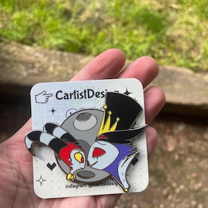 May include: A black and white enamel pin featuring two cartoon characters, one with a red and yellow head and the other with a blue and purple head, kissing on a gray moon. The pin has a white background with black stars and hearts. The text "CarlistDesig" and "instagram: @carlistdesig" are printed on the pin.