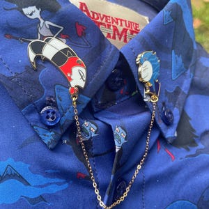 May include: Two gold chains with enamel pins attached to them. The pins are of Finn and Marceline from the cartoon Adventure Time. The chains are on a blue shirt with a pattern of Adventure Time characters.
