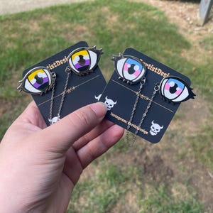 May include: Eye-shaped enamel pins with non-binary and transgender pride flag designs. Each pin has a silver chain and is attached to a black card with the text "artistDesign" and a skull graphic.