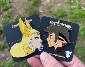 Puede incluir: Dos pines de esmalte con personajes de la serie animada She-Ra y las Princesas del Poder. Los pines están sobre cartulina negra con el texto "Carti Design" y un nombre de usuario de Instagram. Los pines son de Adora y Catra, con Adora en su forma She-Ra y Catra en su forma de gato.
