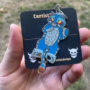 Puede incluir: Pin esmaltado de gato azul con un diseño único, con un gato estilizado con manchas blancas y detalles rojos. El pin está sobre una tarjeta negra con el texto "Cartist" y "artistdesign".
