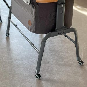 Könnte beinhalten: Ein graues und braunes Babybett mit Metallrahmen und Rädern. Das Babybett hat eine dunkelgraue Stoffaußenseite und einen Holzakzent. Der Rahmen ist dunkelgrau und trägt den Markennamen "MAXI-COSI".