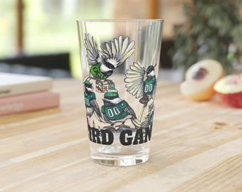 May include: A clear glass pint glass with a design of cartoon birds wearing jerseys and holding a football. The text "Bird Gang" is printed on the bottom of the glass.