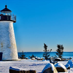 Pode incluir: Um farol branco com telhado e corrimão pretos fica em uma praia coberta de neve com um banco e pedras em primeiro plano. O oceano é visível ao fundo.