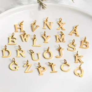 Könnte beinhalten: Eine Sammlung goldfarbener Alphabetanhänger, jeweils mit einer kleinen Schlaufe zum Befestigen an einer Halskette oder einem Armband. Die Buchstaben sind in einer klassischen Serifenschrift gehalten und auf einer weißen Oberfläche angeordnet. Die Anhänger sind gleichmäßig verteilt und gut beleuchtet.
