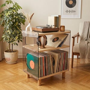 Puede incluir: Un moderno mueble para tocadiscos con laterales de acrílico transparente y estantes de madera. El mueble contiene un tocadiscos, discos de vinilo y auriculares. El mueble está sobre patas de madera.