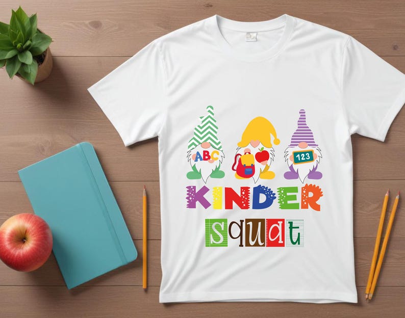 Puede incluir: Camiseta blanca con un gr&aacute;fico de tres gnomos con sombreros y sosteniendo carteles que dicen "ABC", "123" y una mochila. El texto "Kinder Squat" est&aacute; impreso debajo de los gnomos.