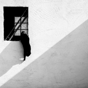 Das Foto zeigt eine schwarze Katze, die aus einem Fenster springt. Die Szene ist in kontrastreichem Schwarz-Weiß gehalten, mit einem scharfen diagonalen Schatten auf der Wand.
