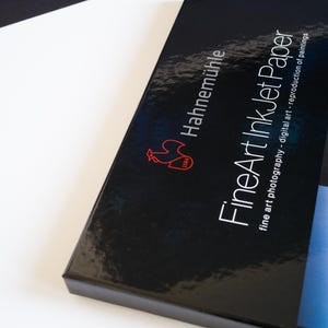 May include: A black rectangular box of Hahnem&uuml;hle FineArt InkJet Paper. The box features a glossy finish, a red rooster logo with the year 1584, and white text that reads FineArt InkJet Paper, fine art photography - digital art - reproduction of paintings.