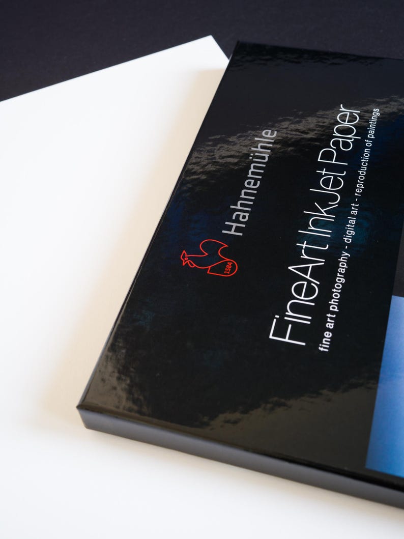 May include: A black rectangular box of Hahnem&uuml;hle FineArt InkJet Paper. The box features a glossy finish, a red rooster logo with the year 1584, and white text that reads FineArt InkJet Paper, fine art photography - digital art - reproduction of paintings.