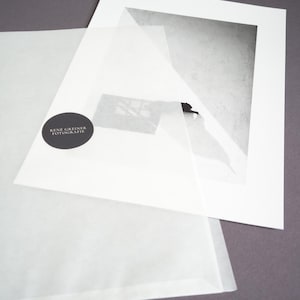 May include: A black and white photographic print mounted on a white mat board, partially covered by a translucent protective sheet featuring a circular black sticker with the white text RENE GREINER FOTOGRAFIE printed in the center.