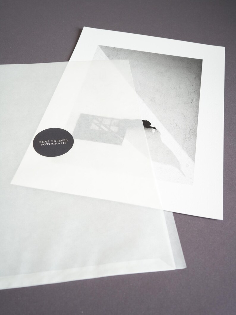May include: A black and white photographic print mounted on a white mat board, partially covered by a translucent protective sheet featuring a circular black sticker with the white text RENE GREINER FOTOGRAFIE printed in the center.