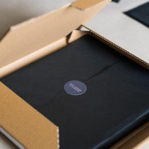 May include: A square cardboard shipping box with an open lid containing a black object wrapped in black paper. A circular dark gray sticker is centered on the paper, featuring the white text RENE GREINER FOTOGRAFIE printed in a clean, sans-serif font.