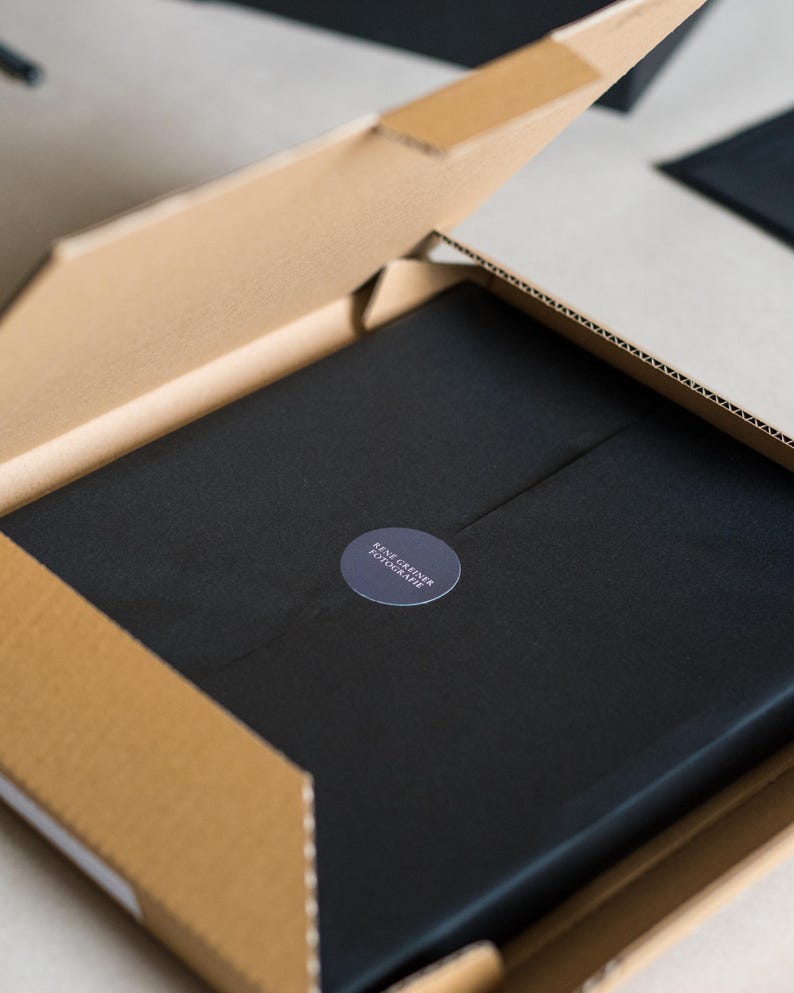 May include: A square cardboard shipping box with an open lid containing a black object wrapped in black paper. A circular dark gray sticker is centered on the paper, featuring the white text RENE GREINER FOTOGRAFIE printed in a clean, sans-serif font.
