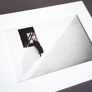 May include: A black and white art print featuring a silhouette of a cat standing on its hind legs, looking out of a rectangular window. The composition includes a stark diagonal shadow across the wall, creating a high-contrast scene in a white mat.