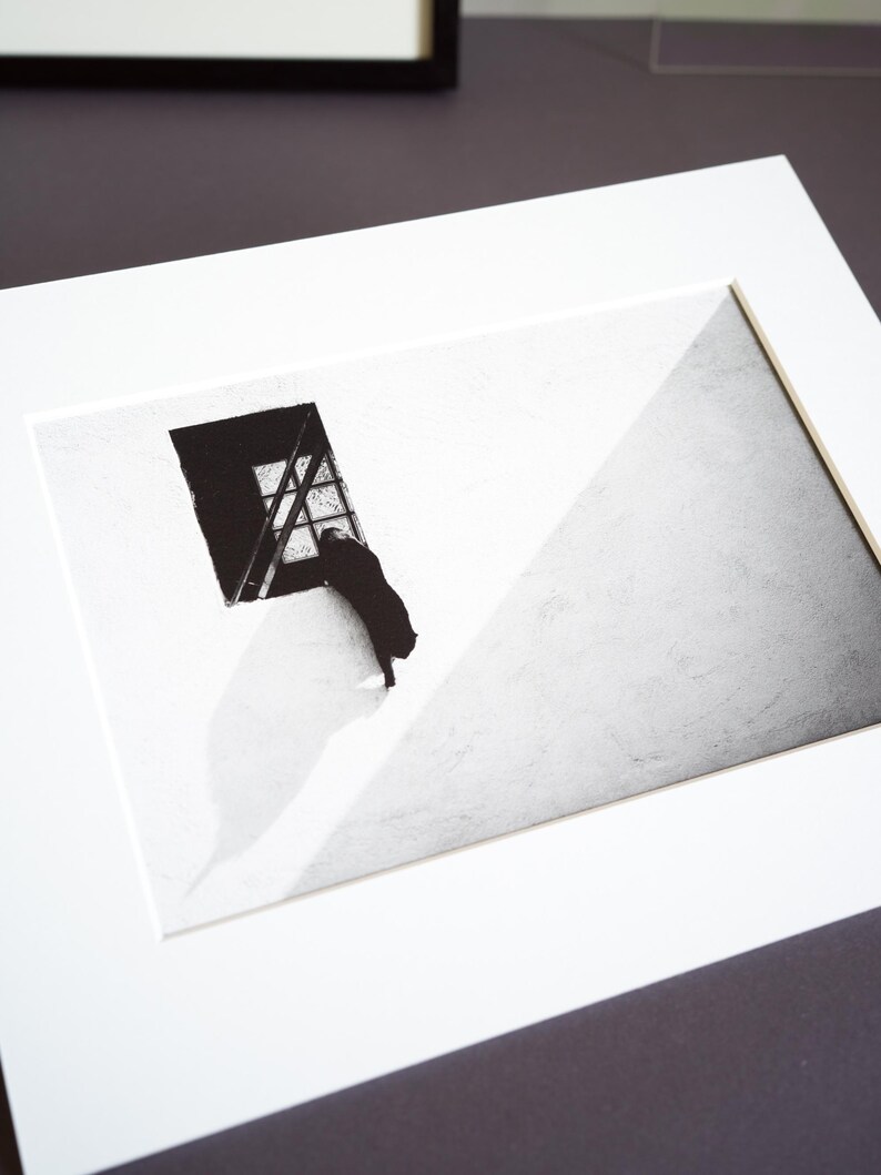 May include: A black and white art print featuring a silhouette of a cat standing on its hind legs, looking out of a rectangular window. The composition includes a stark diagonal shadow across the wall, creating a high-contrast scene in a white mat.