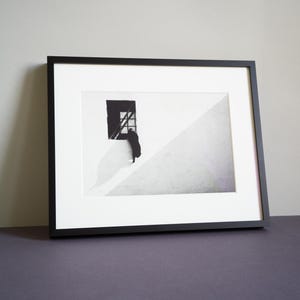 Schwarzweißfotografie, die eine aus einem Fenster springende Katze zeigt. Das Foto ist sehr minimalistisch, mit hohem Kontrast und geometrischen Formen. Die Katze hat ein schwarzes Fell und ist gerade dabei, aus dem Fenster zu springen. Eine Schattenlinie an der Hauswand teilt das Bild diagonal in zwei Hälften.