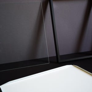 May include: A rectangular black picture frame, a clear acrylic sheet, and a white backing board. The frame is empty, featuring a thin black border. The clear acrylic sheet is positioned upright next to the frame, and the white board lies flat.