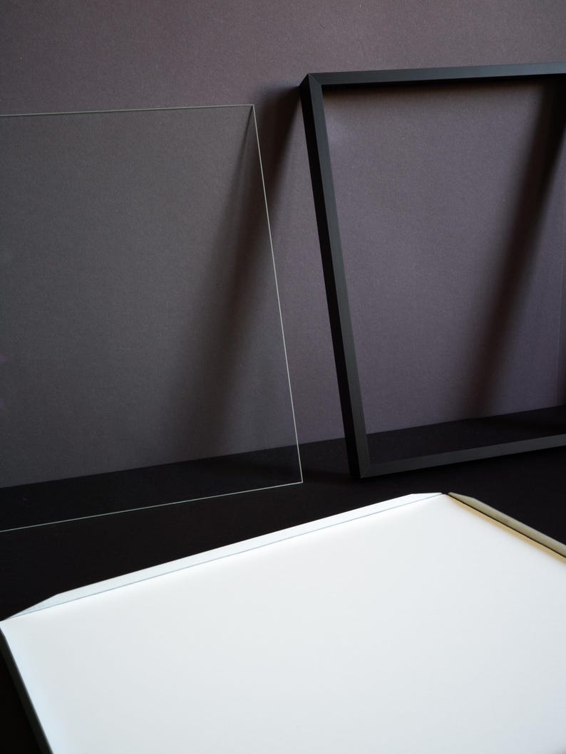 May include: A rectangular black picture frame, a clear acrylic sheet, and a white backing board. The frame is empty, featuring a thin black border. The clear acrylic sheet is positioned upright next to the frame, and the white board lies flat.