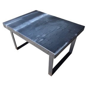 Könnte beinhalten: Ein rechteckiger Couchtisch mit einer dunklen, verkohlten Holzplatte mit sichtbarer Maserung und einem weißen, verwaschenen Finish auf der Oberfläche. Der Tisch wird von vier schwarzen Metallbeinen in einem einfachen Rahmen getragen.