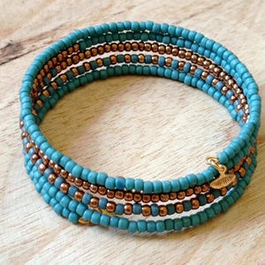 Könnte beinhalten: Ein türkisfarbenes Wickelarmband mit kupferfarbenen Perlen und goldfarbenen Akzenten. Das Armband besteht aus kleinen, runden Perlen und wickelt sich mehrfach um das Handgelenk. Das Armband liegt auf einer hellbraunen Holzoberfläche.