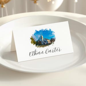 May include: White folded place card with a watercolor image of a church under a blue sky. The name "Ethan Carter" is written in elegant script below the image. The card is displayed on a white plate.