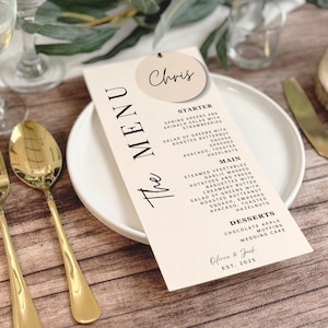 May include: A wedding menu card with the text "The Menu" and a list of dishes. The card is on a white plate with gold silverware. A small tag with the name "Chris" is attached to the menu.