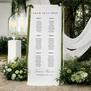 Op de afbeelding: Een witte huwelijkszitting met de tekst "YOUR SEAT Awaits" en gastnamen vermeld onder tabelnummers. De namen staan in zwart script. De namen Jessica + Vivian en de datum 24 september 2026 worden ook weergegeven.