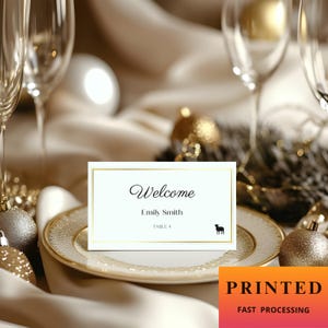 May include: A white place card with gold edging, displaying the word "Welcome" in cursive, the name "Emily Smith", and a table number. A small black dog silhouette is also present. The card is on a decorative plate.
