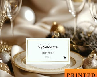 May include: A white place card with gold trim, featuring the word "Welcome" in elegant script, the name "Emily Smith", and a table number. A small black silhouette of a dog is also present. The card is set on a decorative plate.