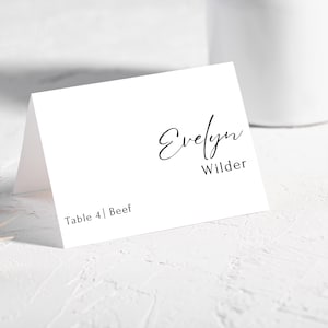 Peut inclure: Une carte de place blanche avec le nom "Evelyn Wilder" en écriture noire élégante. La carte comprend également "Table 4 | Beef" dans une police plus petite. La carte est pliée et posée sur une surface blanche.