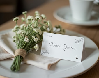 May include: A white place setting with a folded napkin, a small bouquet of white flowers tied with twine, and a place card. The place card reads "Shawn Garcia" in script, with a small illustration of a bird.