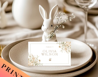 May include: A white ceramic bunny figurine holding delicate white flowers, placed on a layered plate setting. A printed place card with floral accents and the name "Olivia Wilson" is in front of the bunny. The word "PRINTED" is visible.