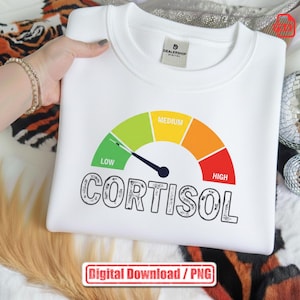 May include: White sweatshirt featuring a cortisol meter graphic. The meter displays levels from green (low) to red (high), with the word "CORTISOL" underneath. The sweatshirt is held by a person.
