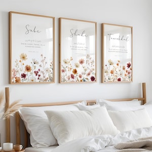 May include: Three framed wall art pieces with floral designs and calligraphy. The frames are light brown, and the artwork features a variety of flowers in shades of red, yellow, and white. The calligraphy is in black. The art is displayed above a bed with white bedding.