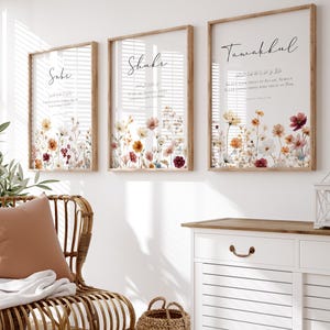 May include: Three framed floral art prints with Arabic calligraphy. The prints feature a watercolor floral design in shades of yellow, orange, and red. The frames are a light brown color, and the prints are displayed on a white wall.