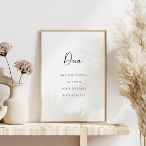 K&ouml;nnte beinhalten: Ein gerahmter Druck mit dem Text "Dua has the power to turn your dreams into reality" auf wei&szlig;em Hintergrund. Der Druck steht auf einem Holzregal mit zwei Vasen und einer getrockneten Pampasgras-Pflanze.